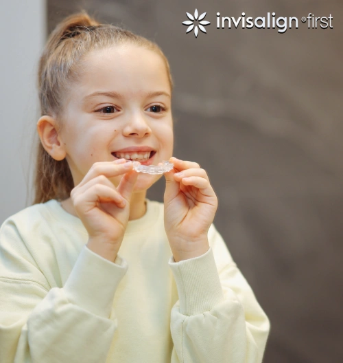Young girl holding Invisalign clear aligners, smiling in clinical orthodontic setting.